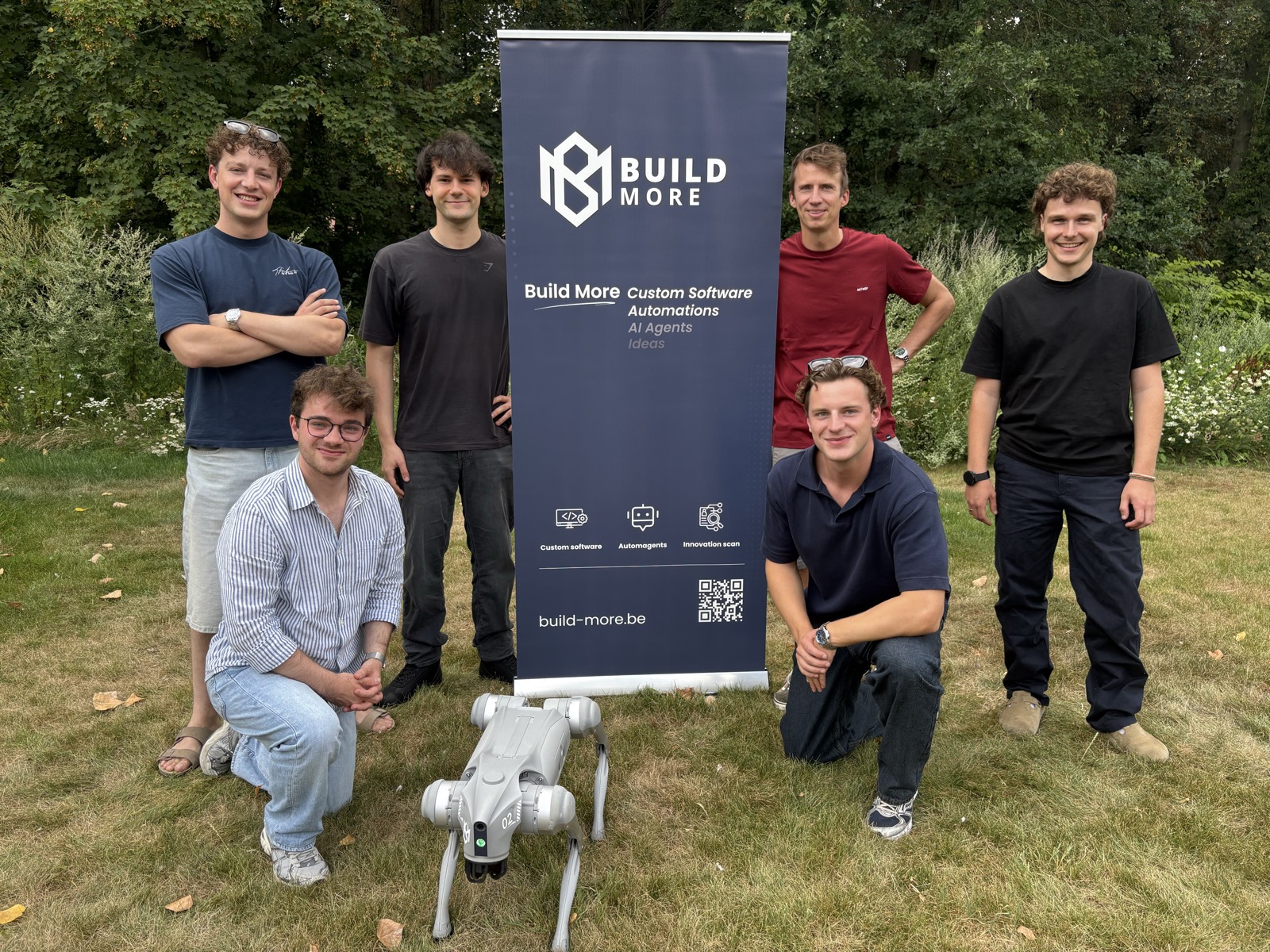 Het team van Build More bij de eigen banner, met een robothond op het gras.